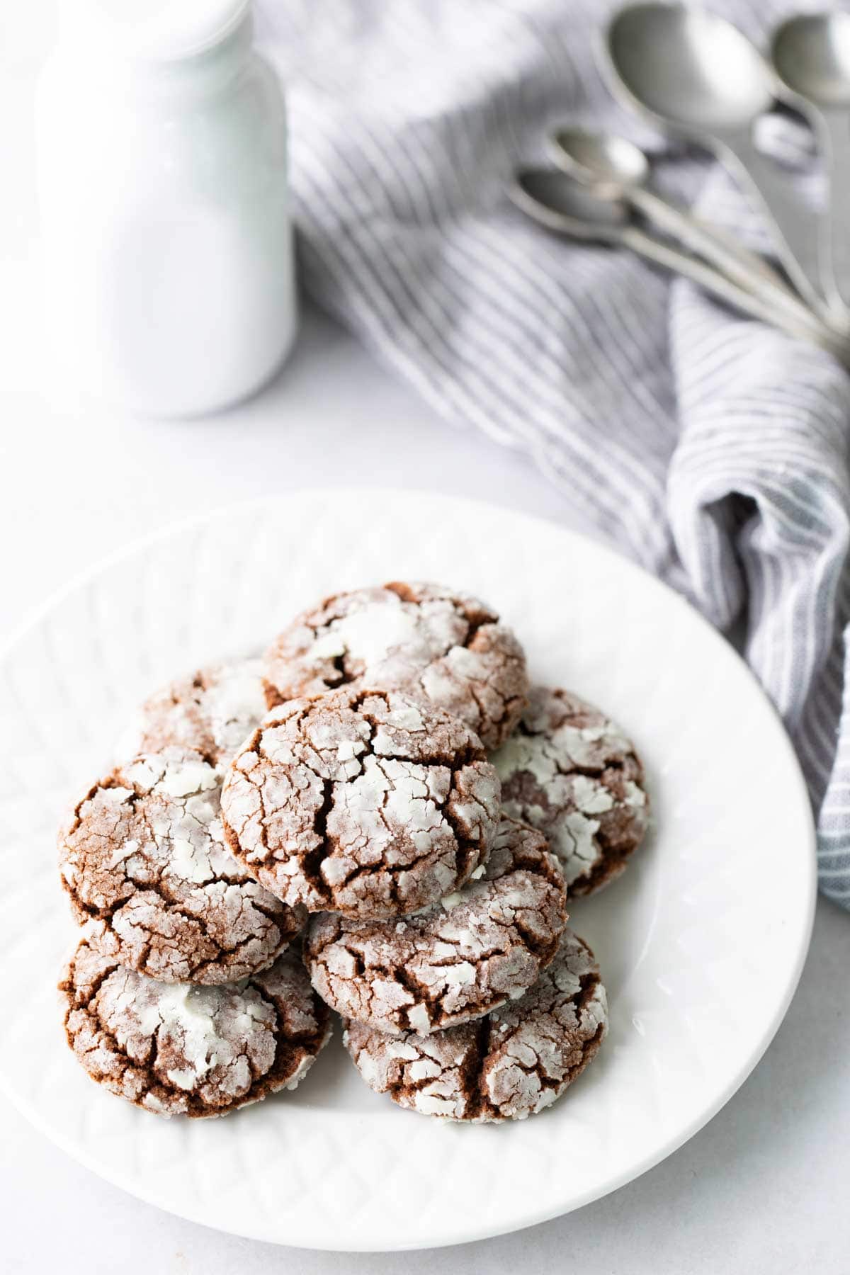 A white plate with a stack of chocolate crinkle cookies, dusted with powdered sugar, sits on a white surface near a striped cloth and metal measuring spoons.
