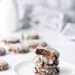 A stack of three chocolate crinkle cookies on a white plate, with the top cookie partially eaten; more cookies and a milk bottle are in the blurred background.