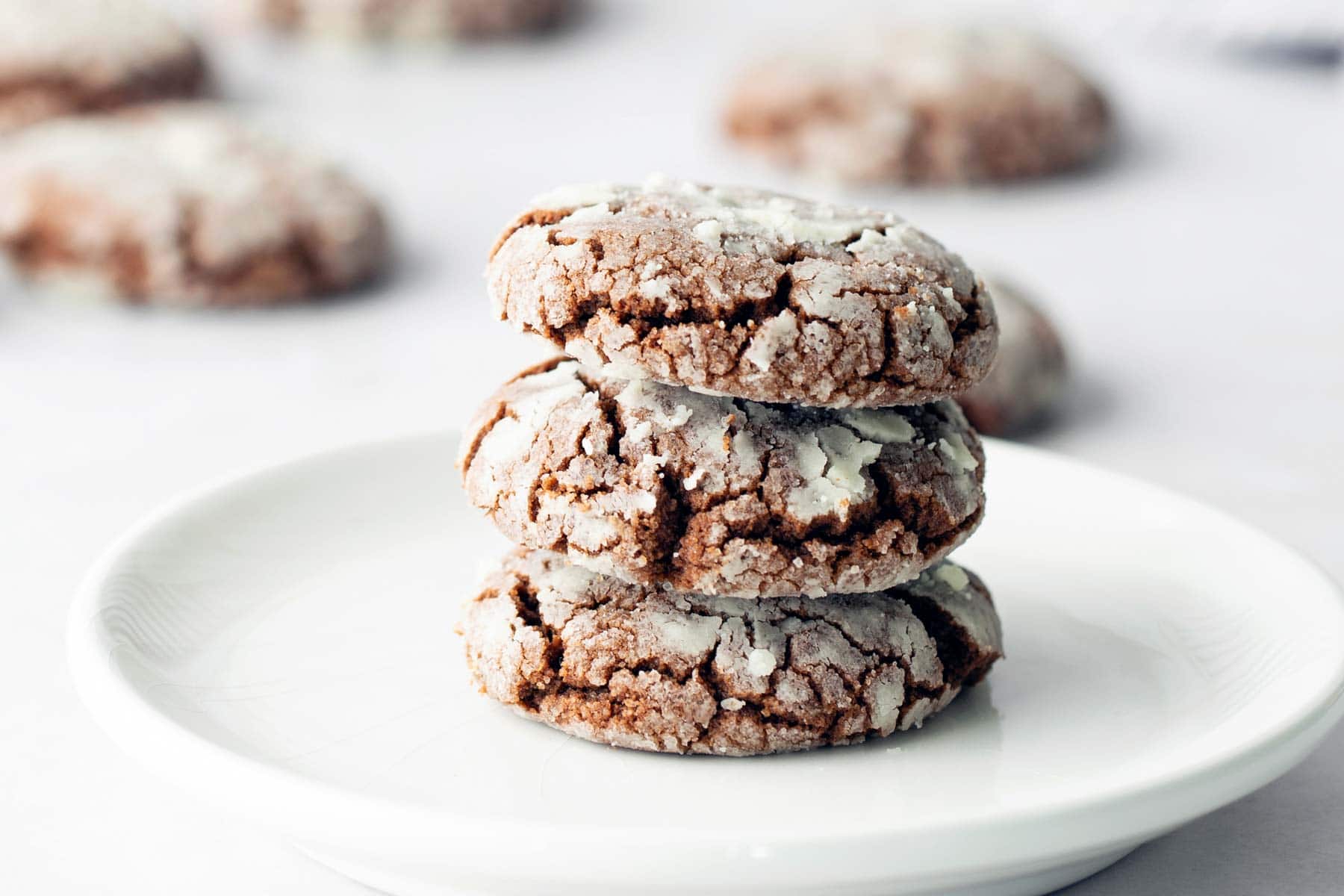 Three chocolate crinkle cookies stacked on a white plate, with more cookies blurred in the background.