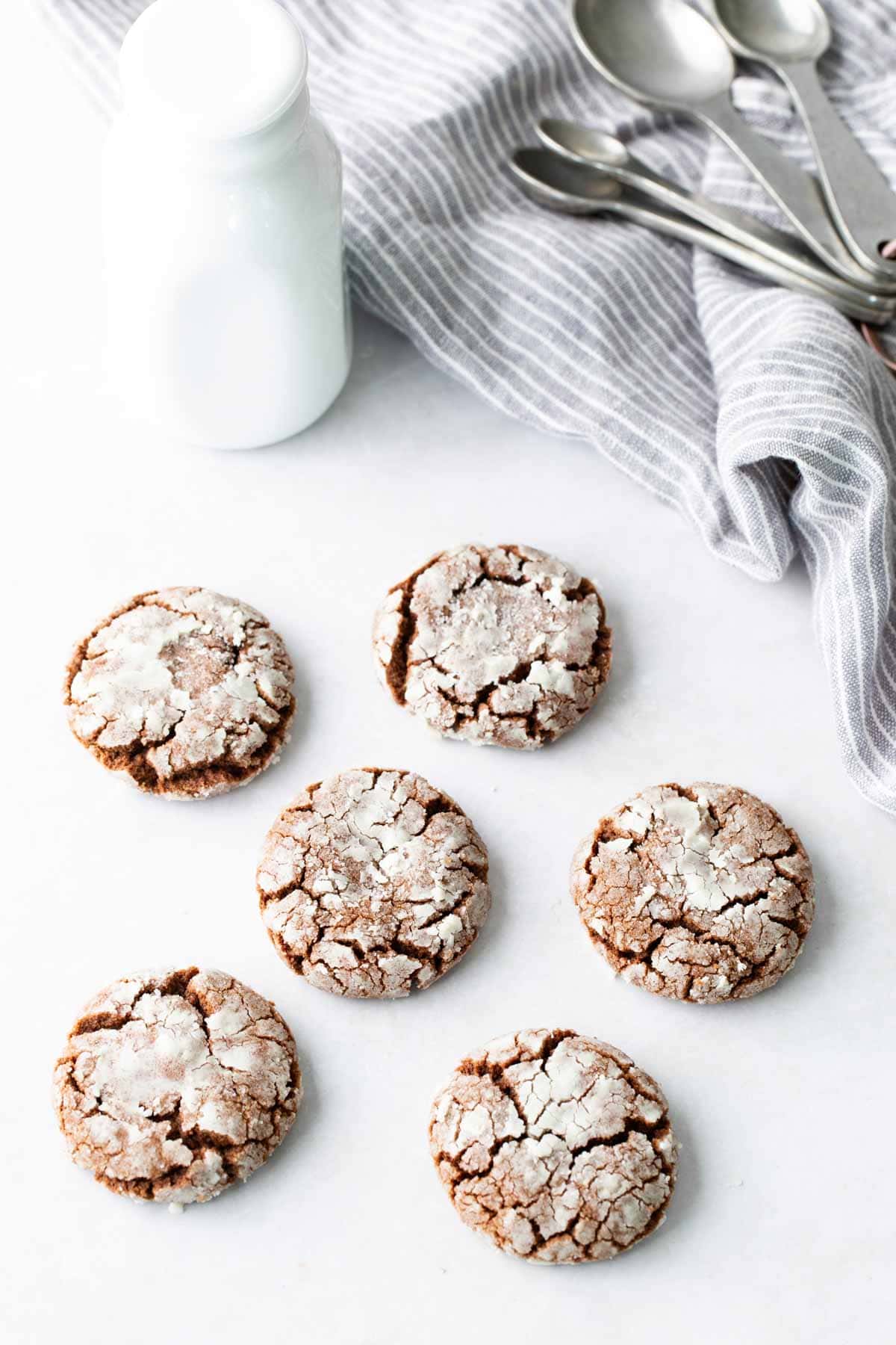 Six chocolate crinkle cookies are arranged on a white surface next to a bottle of milk, a gray striped cloth, and a stack of metal spoons.