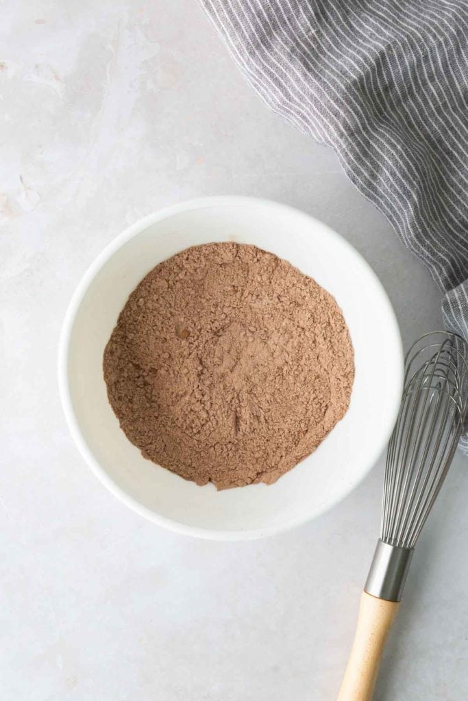 A white bowl filled with cocoa powder sits on a light countertop next to a metal whisk and a gray striped cloth.