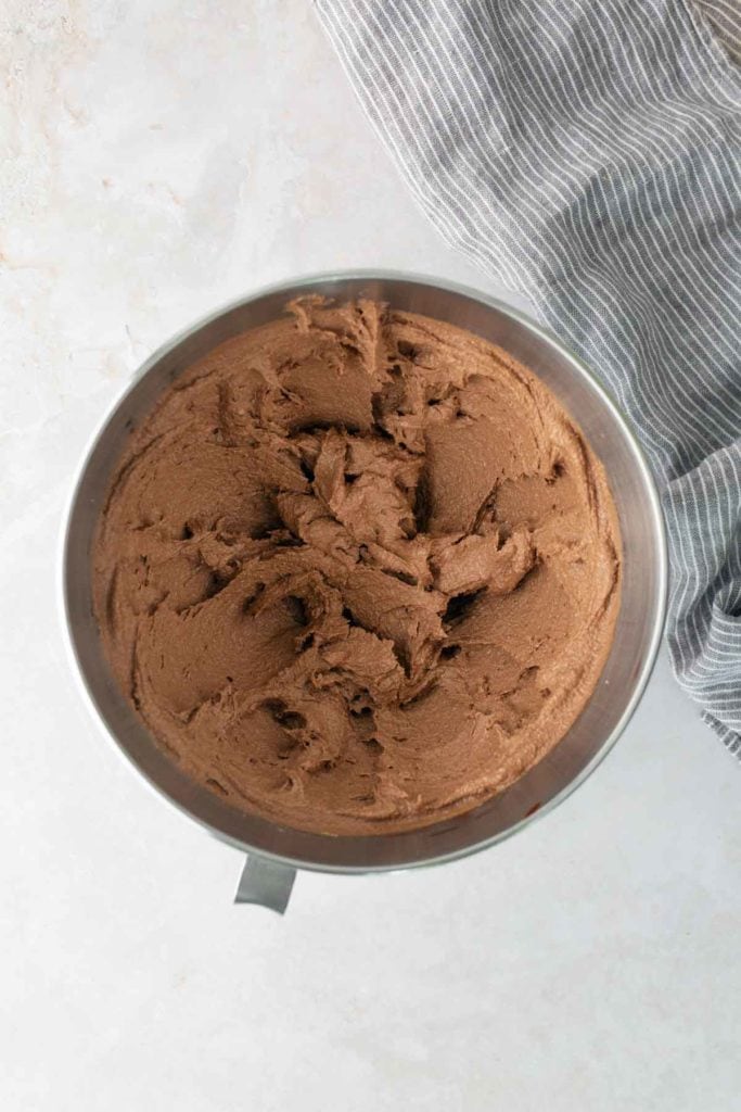 A metal mixing bowl filled with whipped chocolate frosting sits on a light surface next to a gray and white striped cloth.