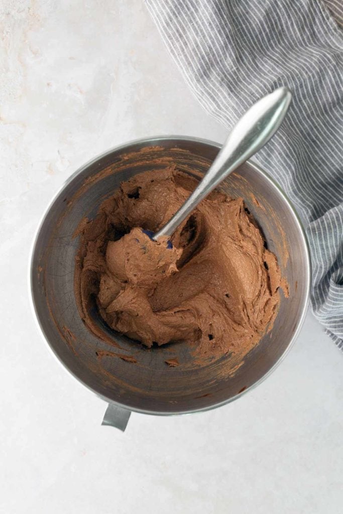 A metal mixing bowl with chocolate frosting and a spoon inside, placed on a light surface next to a gray striped cloth.