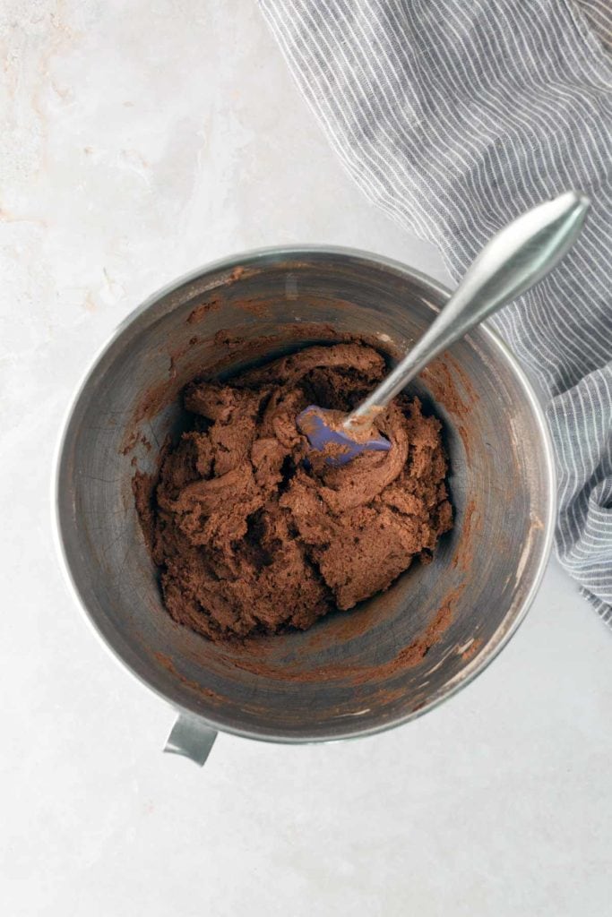 A mixing bowl filled with chocolate cookie dough and a metal spoon, placed on a light surface next to a striped cloth.