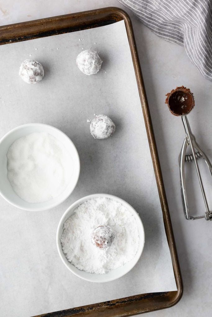 A baking sheet lined with parchment paper holds three chocolate dough balls dusted in powdered sugar, next to two bowls of sugar and a metal cookie scoop.