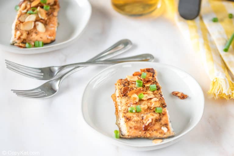 A slice of cooked salmon topped with sliced almonds and chopped green onions is served on a white plate, with two forks beside it.