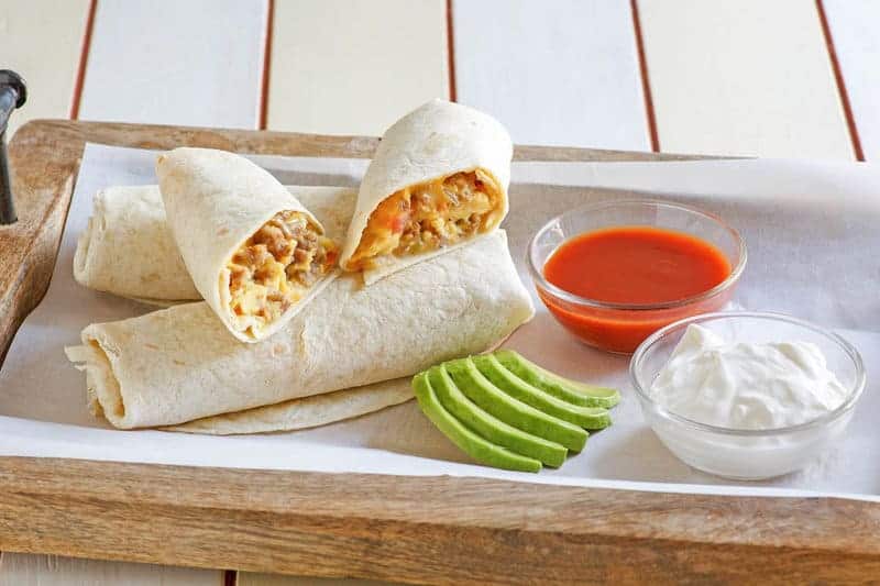 A wooden tray with breakfast burritos, sliced avocado, a bowl of red sauce, and a bowl of sour cream on parchment paper.