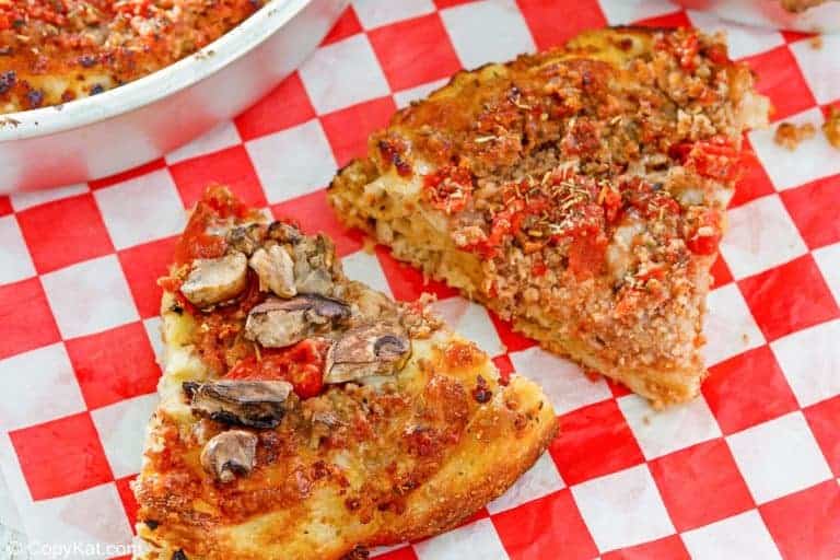 Two slices of pizza with thick crust on a red and white checkered tablecloth, one topped with mushrooms and vegetables, the other with tomato and herbs.