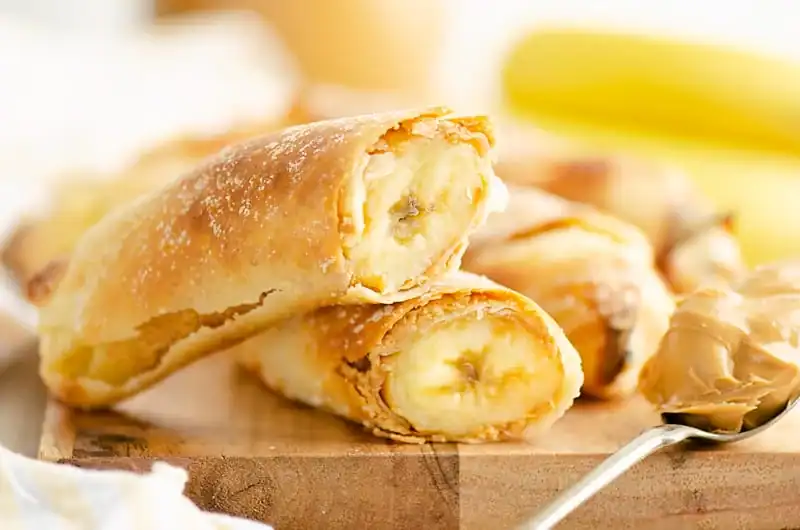 Close-up of two banana-filled pastry rolls stacked on a wooden board, with a spoonful of peanut butter and a blurred banana in the background.
