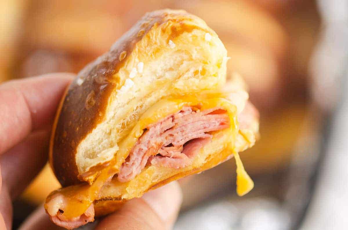 A hand holds a bitten sandwich with layers of sliced meat and melted cheese inside a soft, golden-brown bread roll.