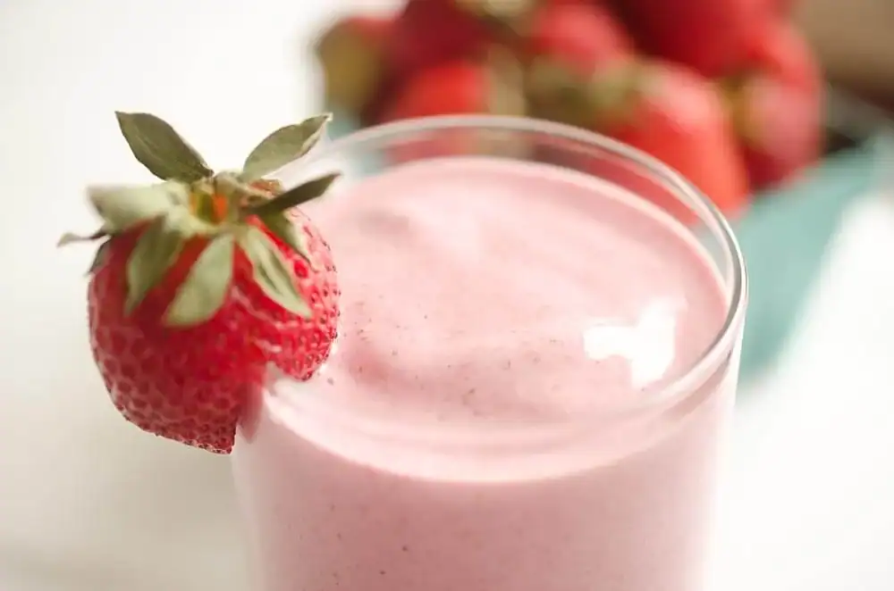 A glass of pink strawberry smoothie with a whole strawberry garnishing the rim. Blurred strawberries are visible in the background.