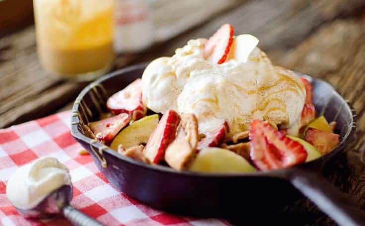 A cast iron skillet filled with sliced apples, strawberries, and a large scoop of ice cream sits on a red and white checkered cloth.