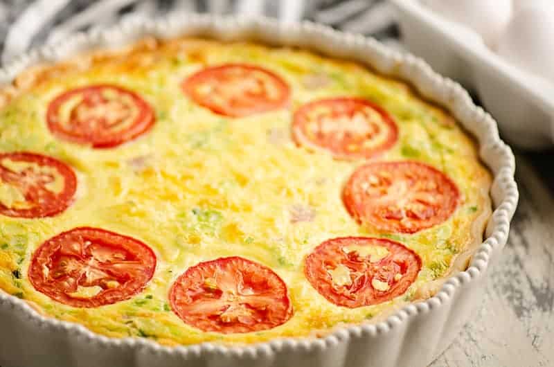A baked quiche in a white fluted dish, topped with sliced tomatoes and a golden, cheesy surface.
