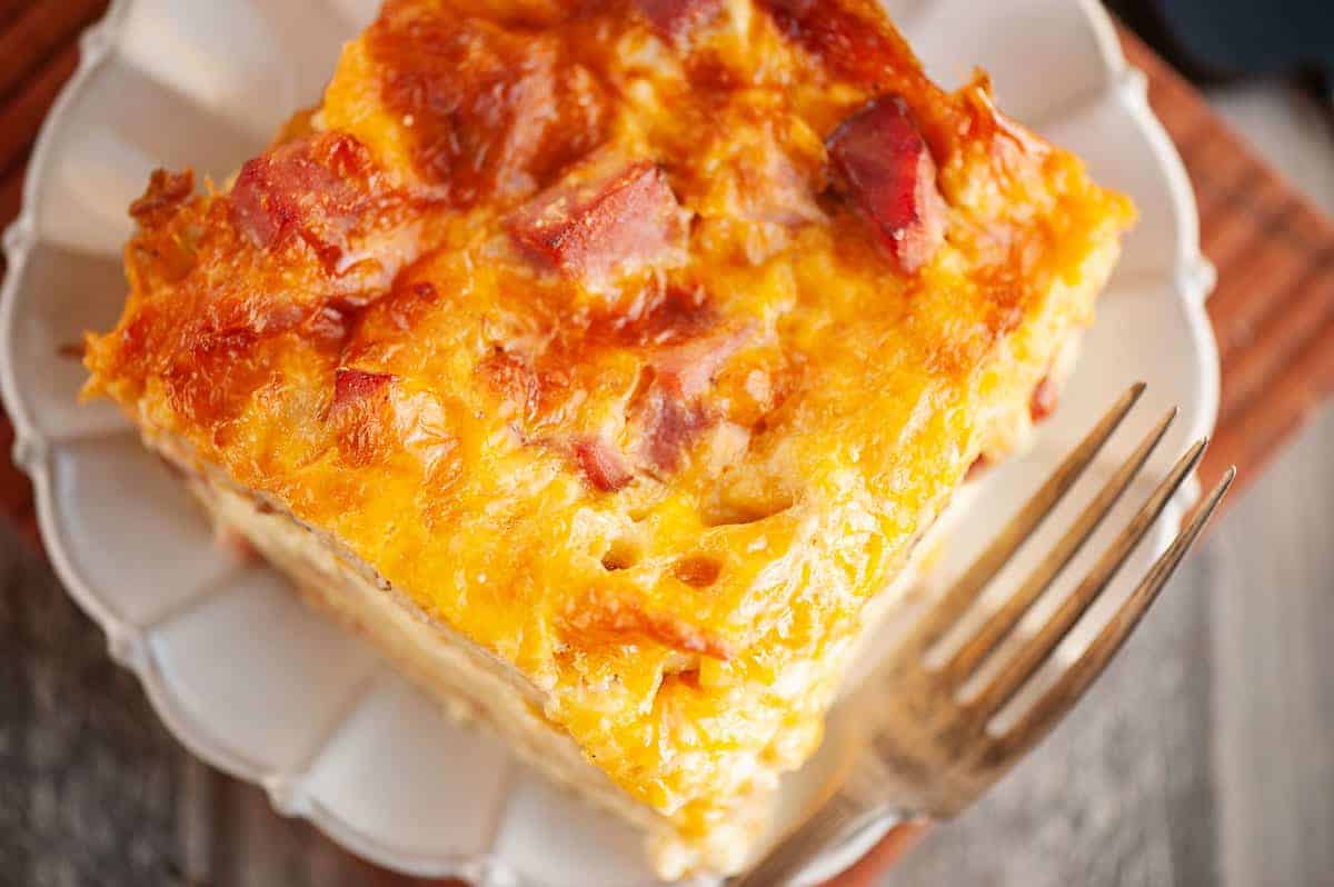 A close-up of a square slice of baked casserole with melted cheese and pieces of ham on a white plate next to a fork.
