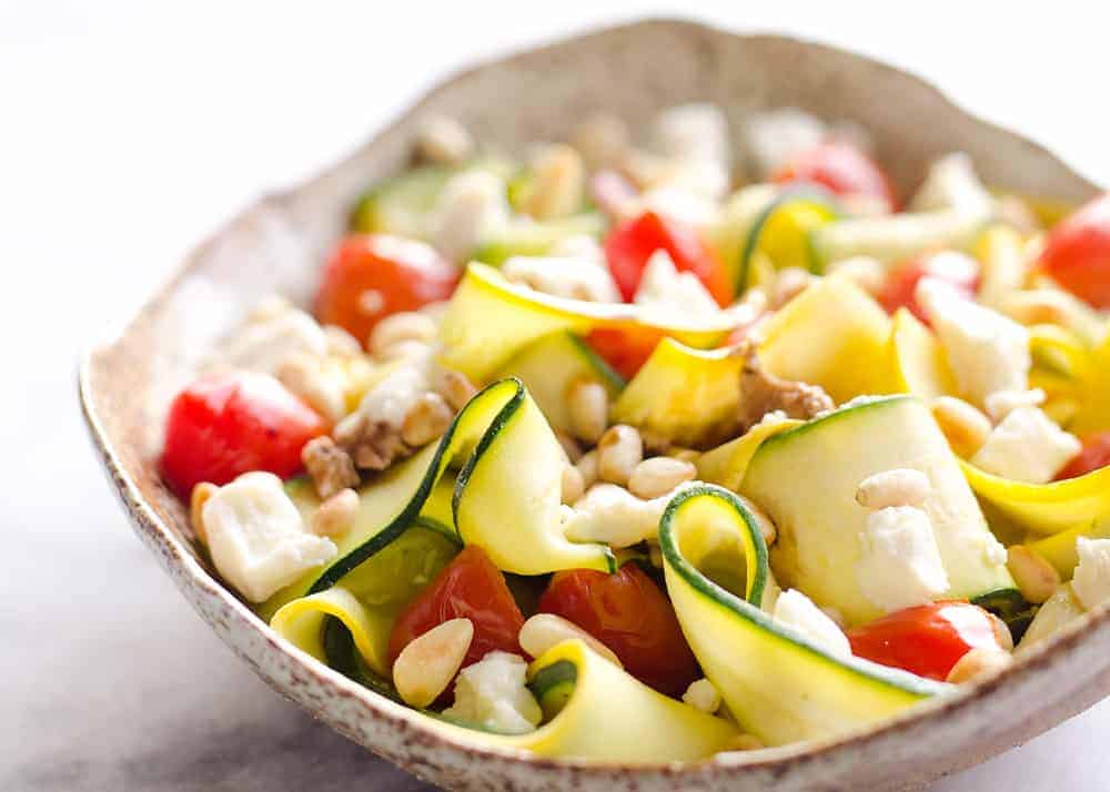 A bowl of zucchini ribbon salad with cherry tomatoes, feta cheese, pine nuts, and walnuts on a light background.