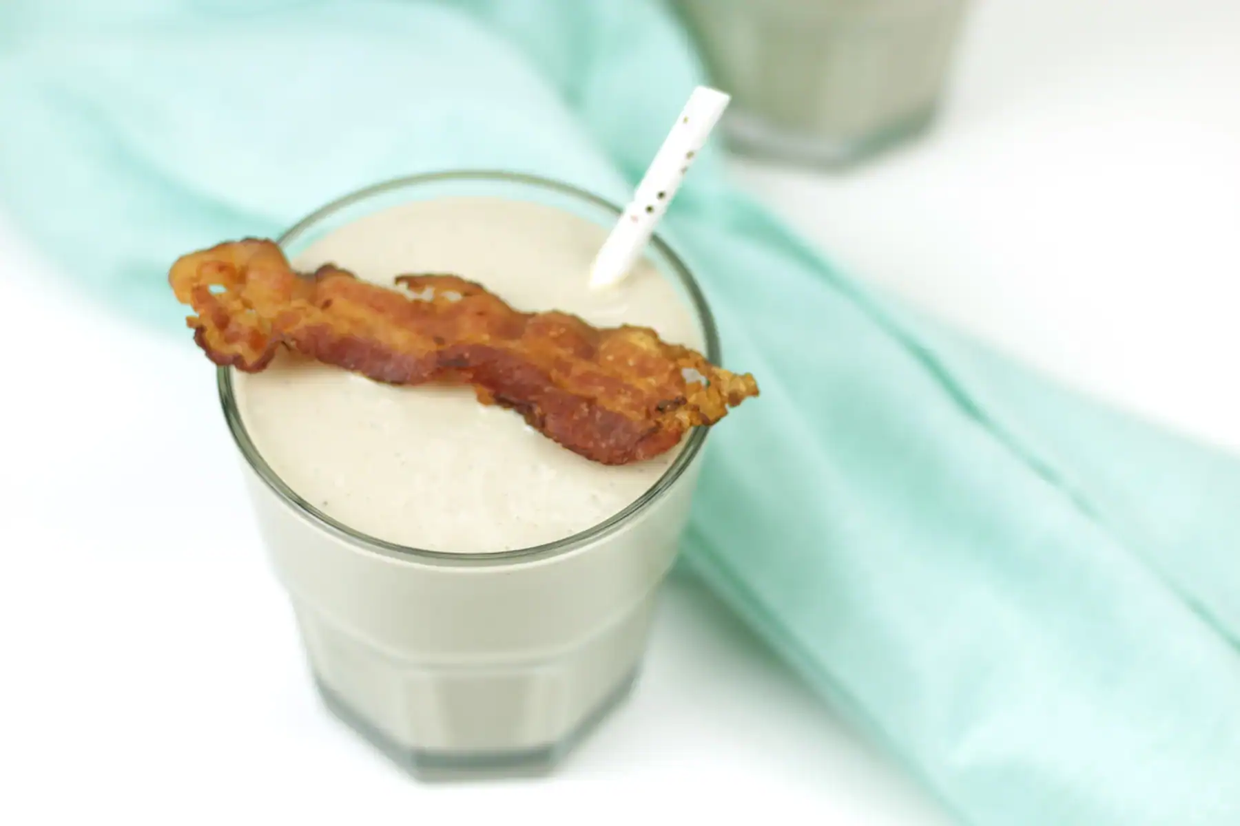 A glass of light-colored milkshake with a paper straw, topped with a strip of crispy bacon, placed next to a light blue cloth.