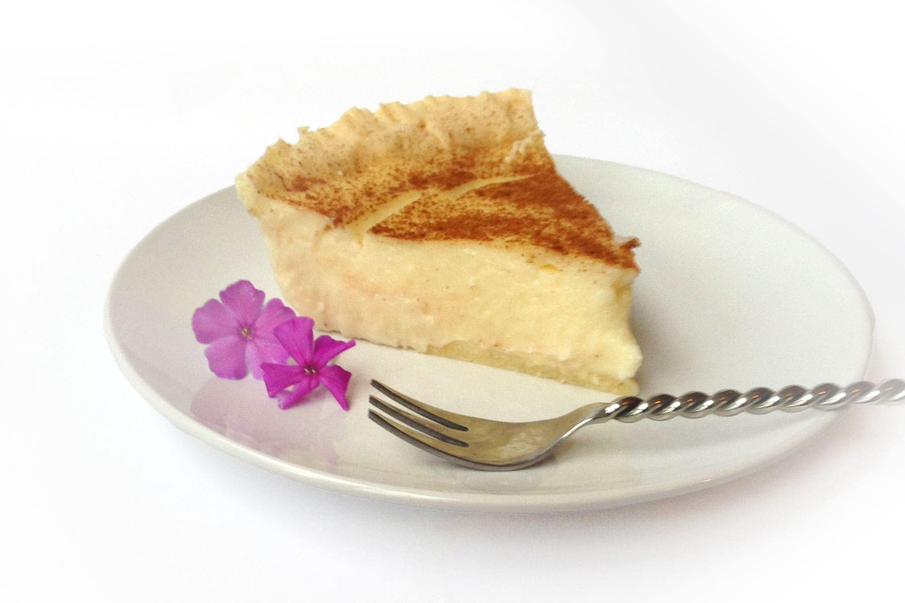 A slice of custard pie on a white plate with a fork and two small purple flowers beside it.