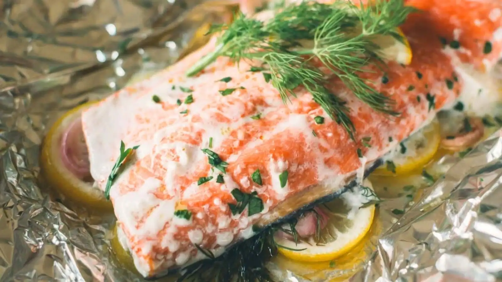 A baked salmon fillet topped with fresh herbs and lemon slices, resting on a sheet of aluminum foil.