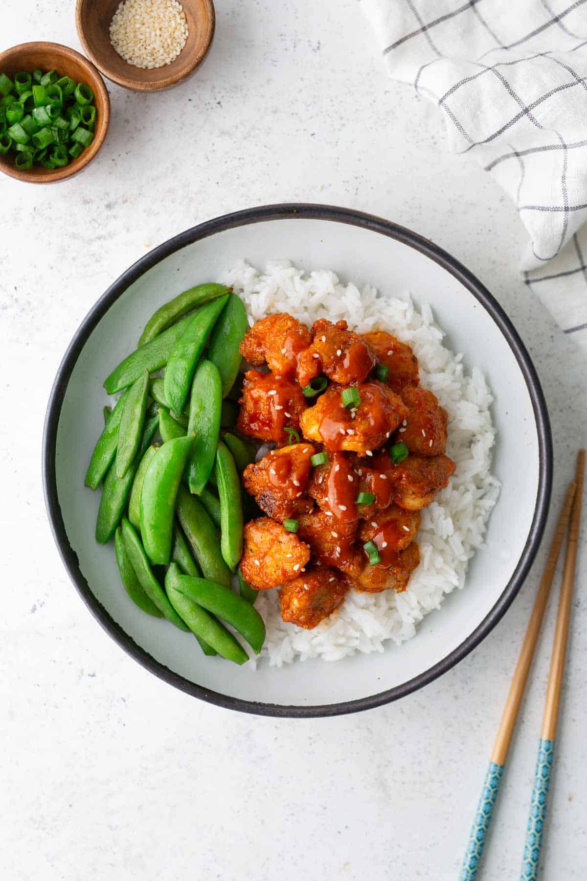 A bowl of white rice topped with saucy cauliflower florets and sliced green onions, served with snap peas. Bowls of sesame seeds and chopped scallions, a cloth, and chopsticks are nearby.