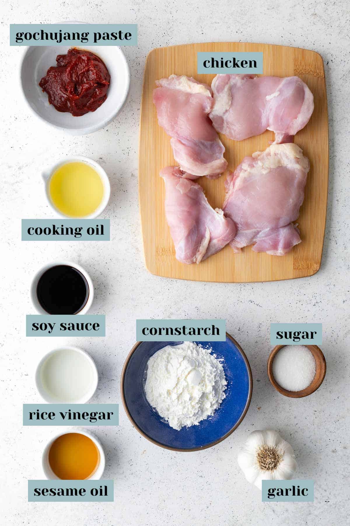Raw chicken thighs on a wooden board with bowls of gochujang paste, cooking oil, soy sauce, rice vinegar, sesame oil, cornstarch, sugar, and garlic on a white surface.