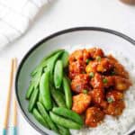 A bowl of white rice topped with glazed chicken pieces, sliced green onions, and snap peas, with chopsticks placed beside the bowl.