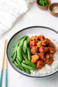 A bowl of white rice topped with glazed chicken pieces, sliced green onions, and snap peas, with chopsticks placed beside the bowl.