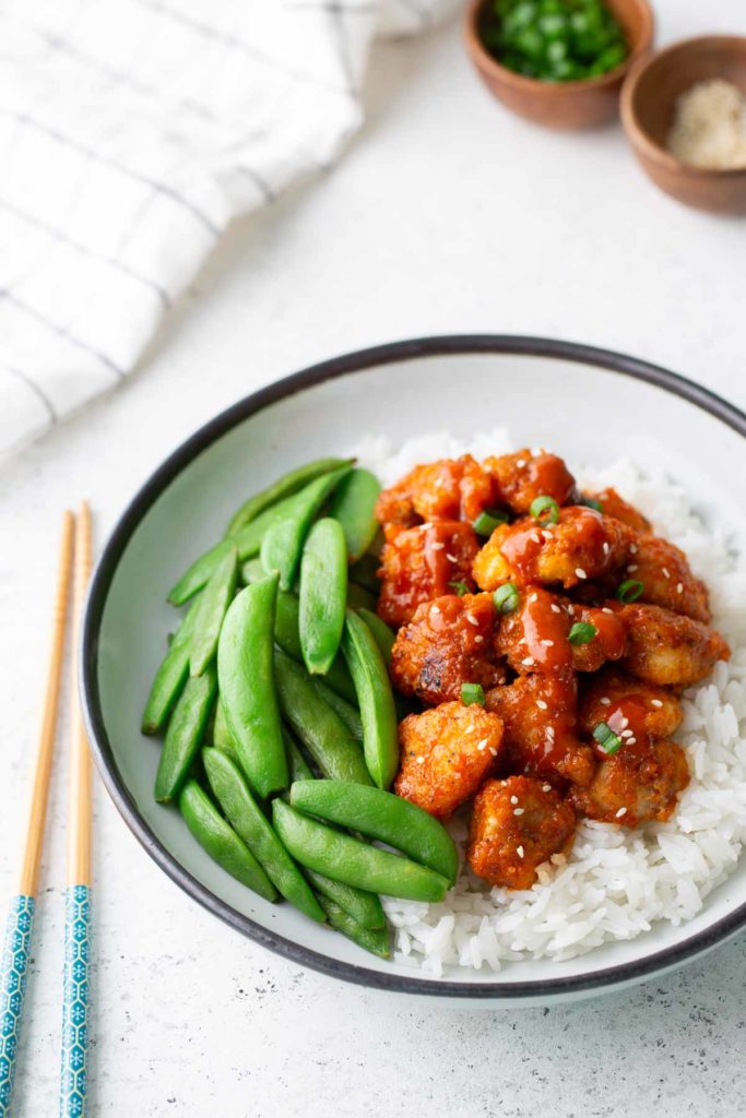 Gochujang Chicken A bowl of white rice topped with glazed chicken pieces, sliced green onions, and snap peas, with chopsticks placed beside the bowl.
