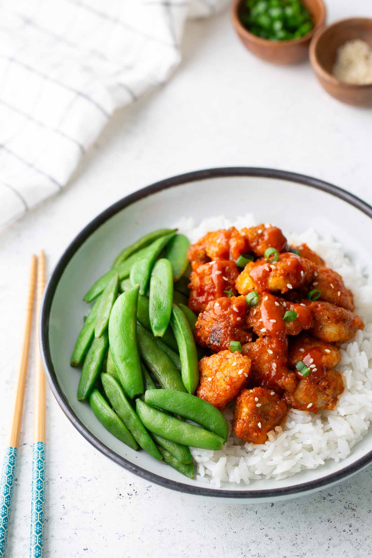A bowl of white rice topped with glazed chicken pieces, sliced green onions, and snap peas, with chopsticks placed beside the bowl.