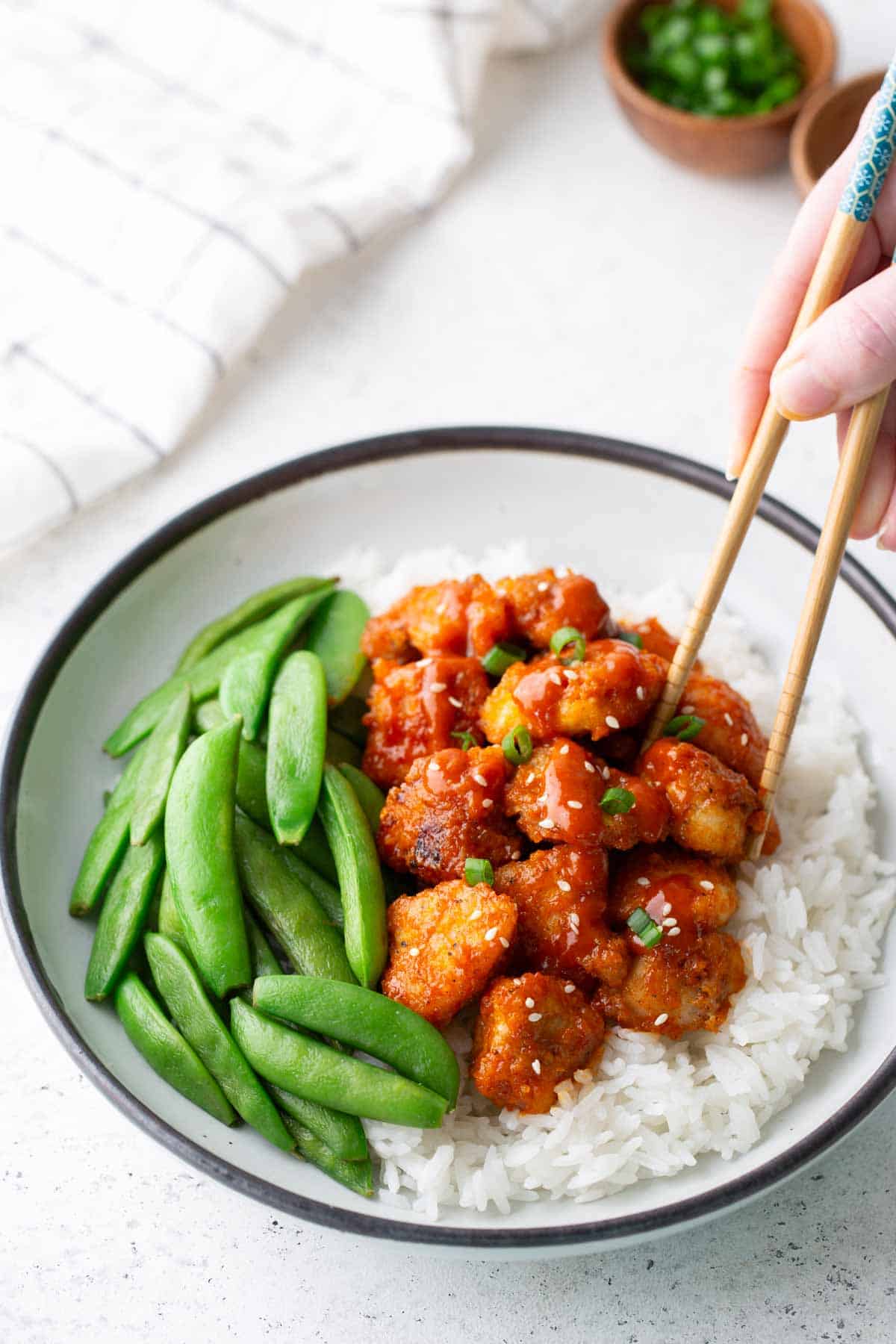 A bowl of white rice topped with glazed chicken pieces, sliced green onions, and snap peas; a hand uses chopsticks to pick up a piece of chicken.