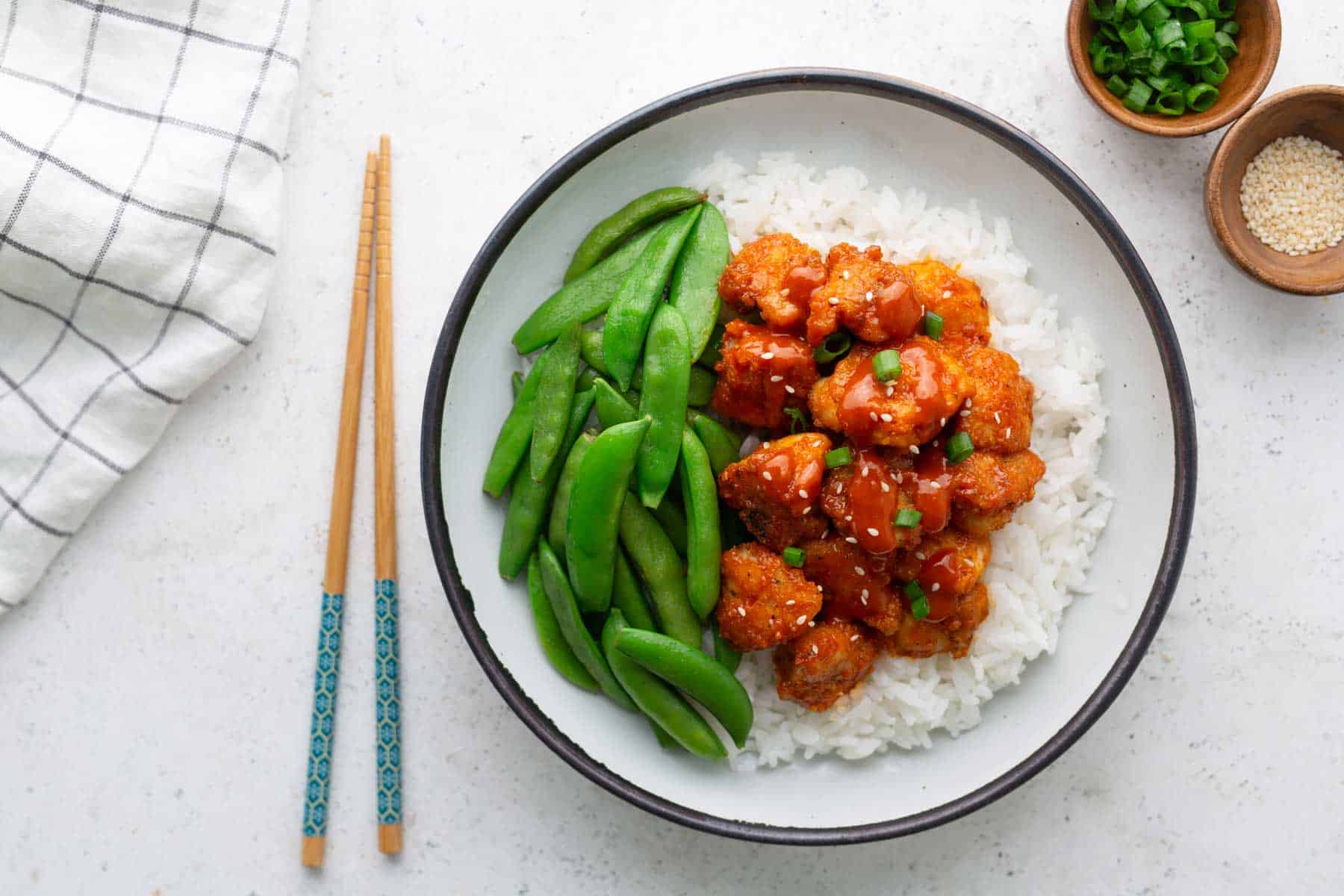 A bowl of white rice topped with glazed chicken pieces and steamed snap peas, with chopsticks, chopped green onions, and sesame seeds on the side.