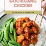 A bowl of white rice topped with gochujang chicken, sliced green onions, and snap peas. Chopsticks are picking up a piece of chicken.