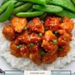 A bowl of white rice topped with gochujang chicken, sliced green onions, sesame seeds, and served with sugar snap peas.