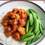 A bowl of white rice topped with gochujang chicken pieces, sliced green onions, sesame seeds, and a side of snap peas. Text at top reads "easy gochujang chicken.