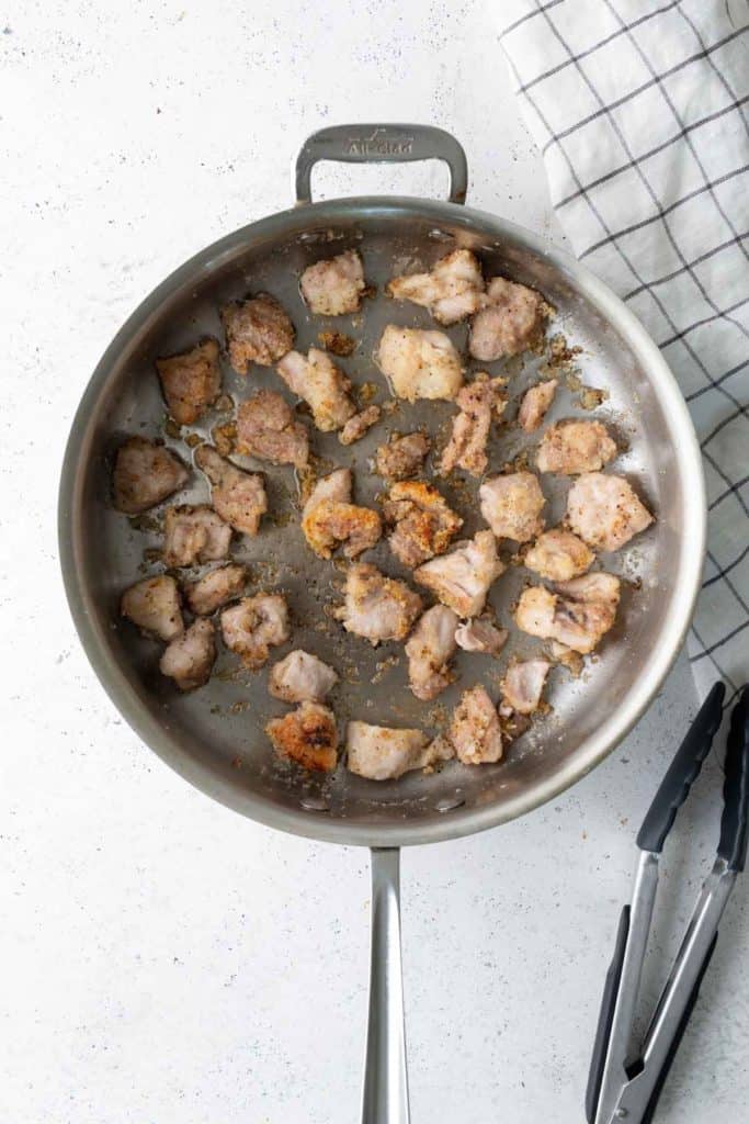 A stainless steel skillet with browned chicken pieces, sitting on a white surface next to metal tongs and a checkered cloth.