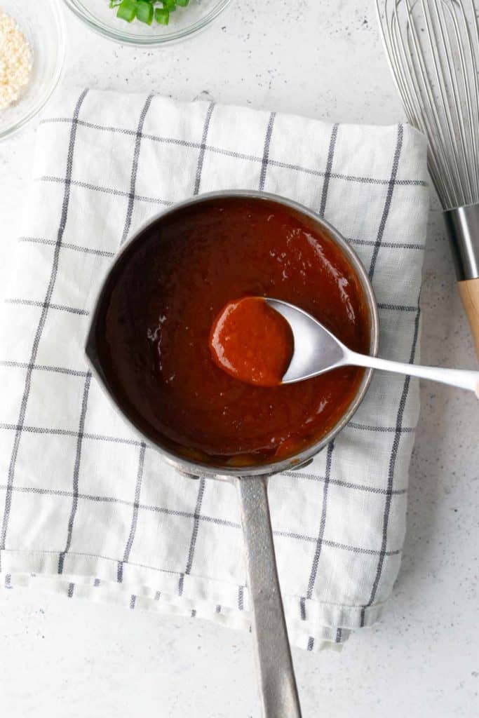 A saucepan filled with a thick red sauce, a spoon resting inside, placed on a white towel with black stripes; a whisk and small bowls are nearby.