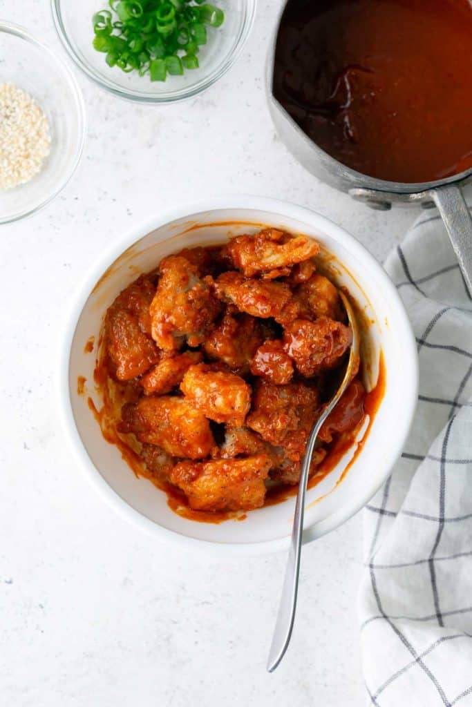 A bowl of sauced fried chicken pieces with a spoon, next to a pot of sauce, chopped green onions, sesame seeds, and a checkered cloth on a white surface.
