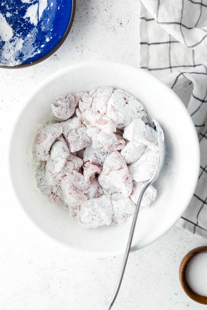 A white bowl containing sliced pieces of raw meat coated in flour, with a metal spoon resting inside. A checkered cloth and a blue bowl with flour are nearby on a white surface.
