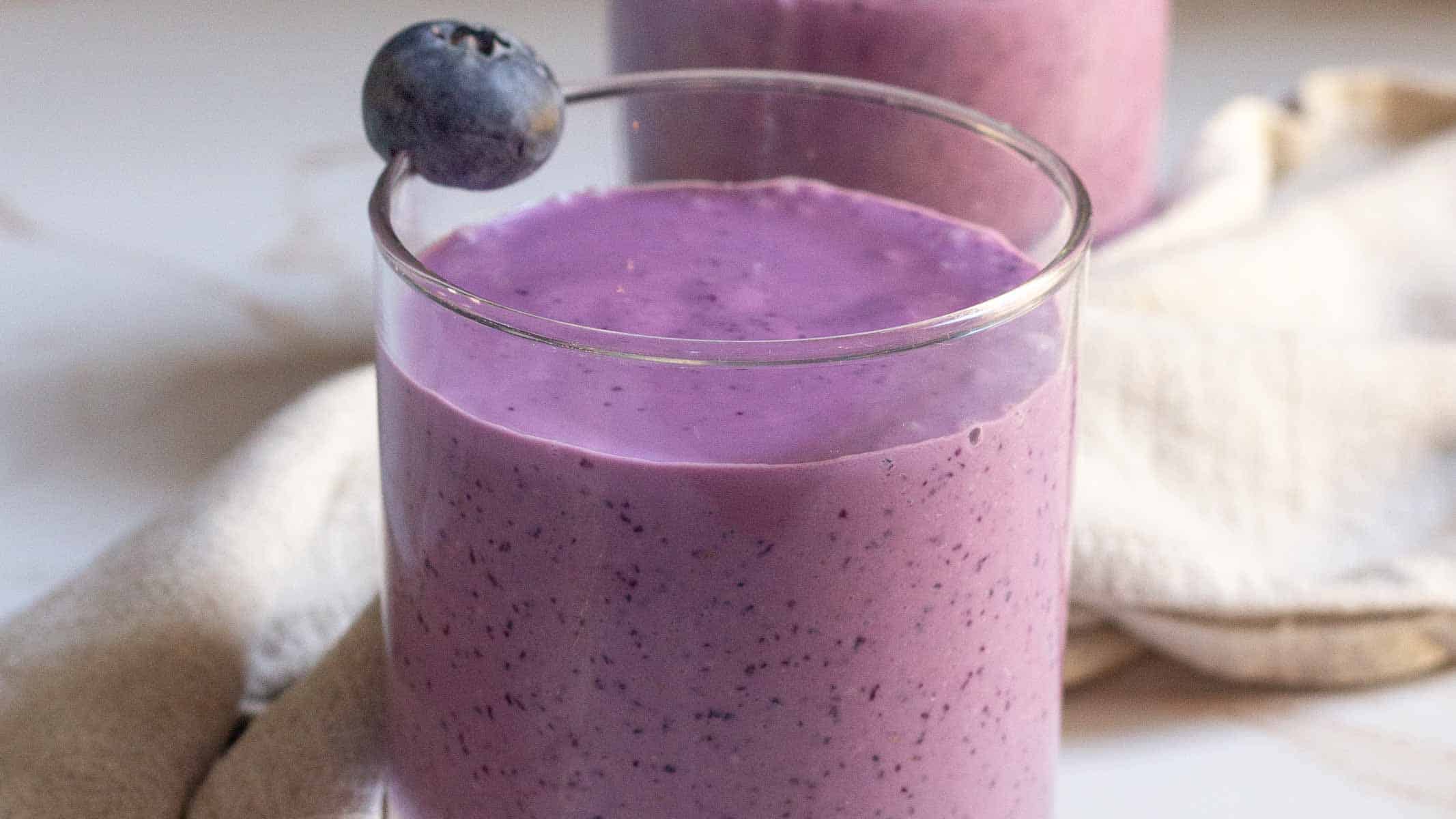 A glass of purple blueberry smoothie with a blueberry garnish on the rim, placed in front of a light-colored cloth.