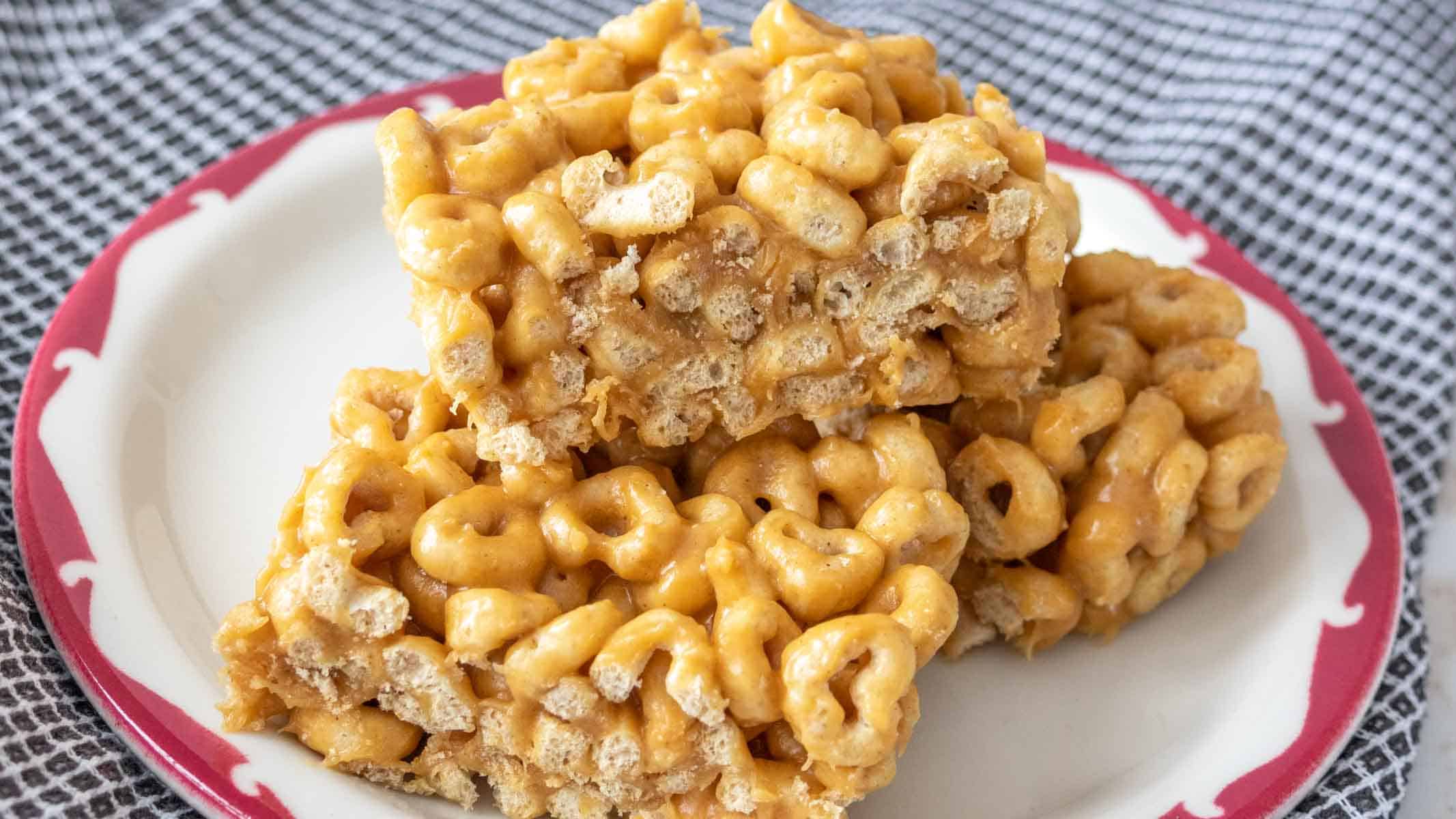 Three peanut butter Cheerio bars are stacked on a white plate with a red border, set on a black-and-white checkered cloth.