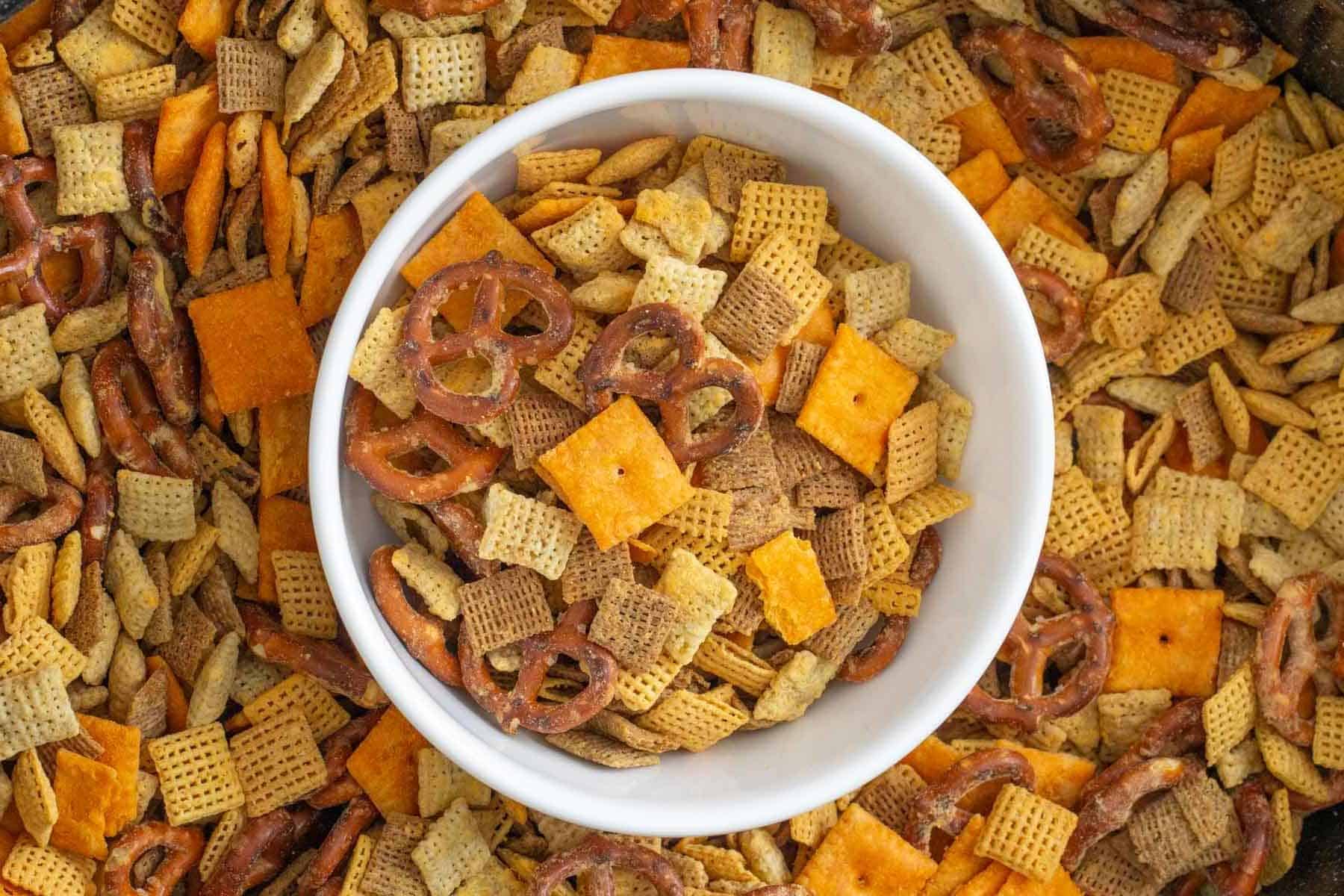 A white bowl filled with a snack mix of pretzels, square crackers, and cereal pieces sits on top of more of the same mix.