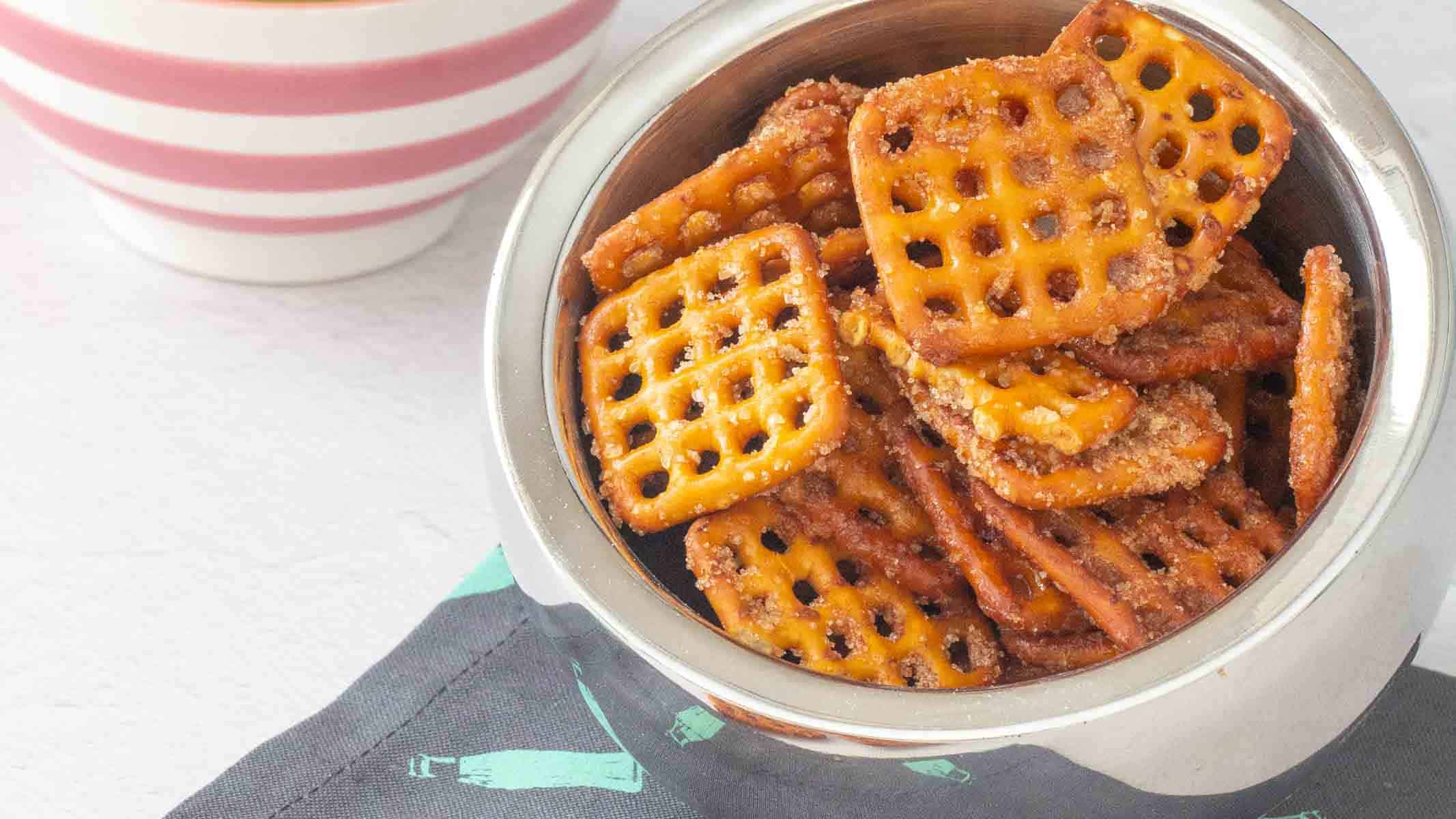 A metal bowl filled with square pretzel crackers coated in seasoning, placed on a dark napkin near a striped ceramic bowl.