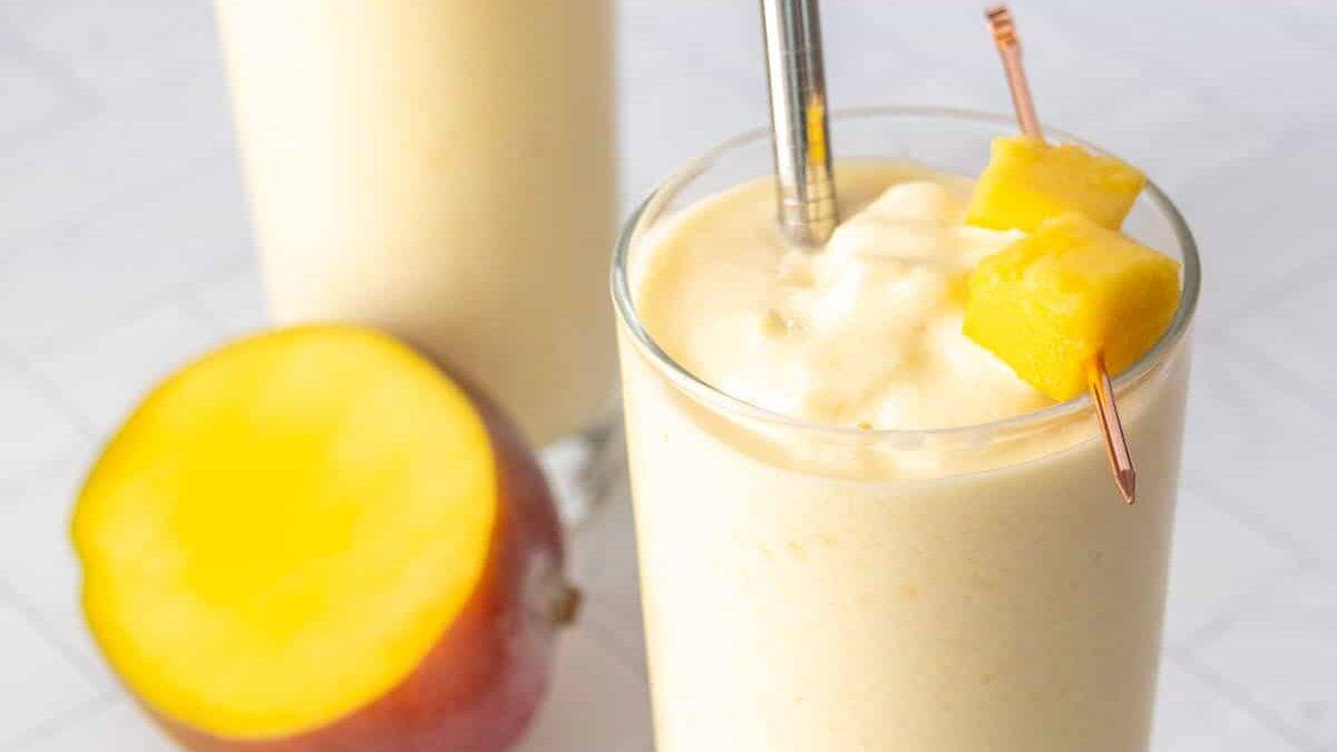 Two glasses of mango smoothie with metal straws, garnished with mango chunks on skewers, next to a halved mango on a white surface.