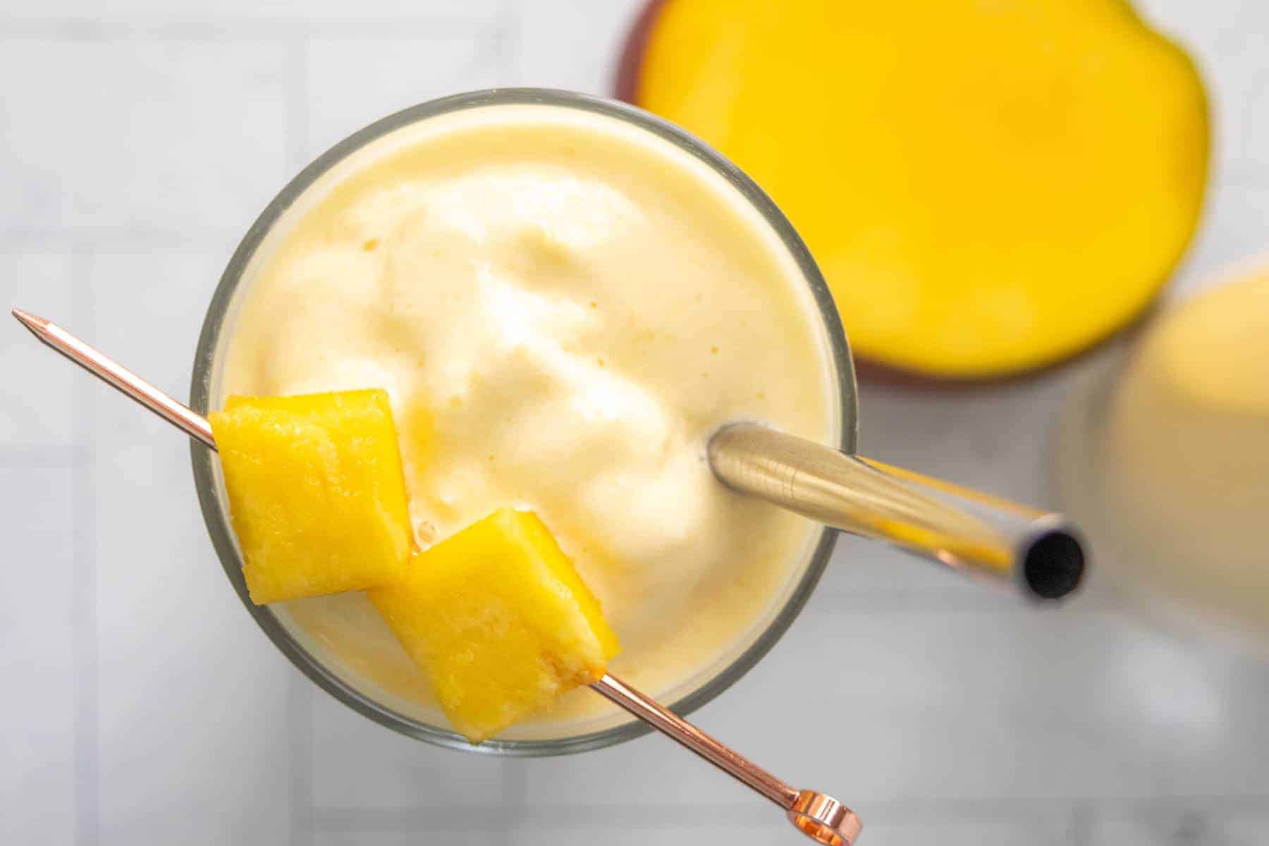 A glass of mango smoothie with two pieces of mango on a cocktail pick and a metal straw, viewed from above.