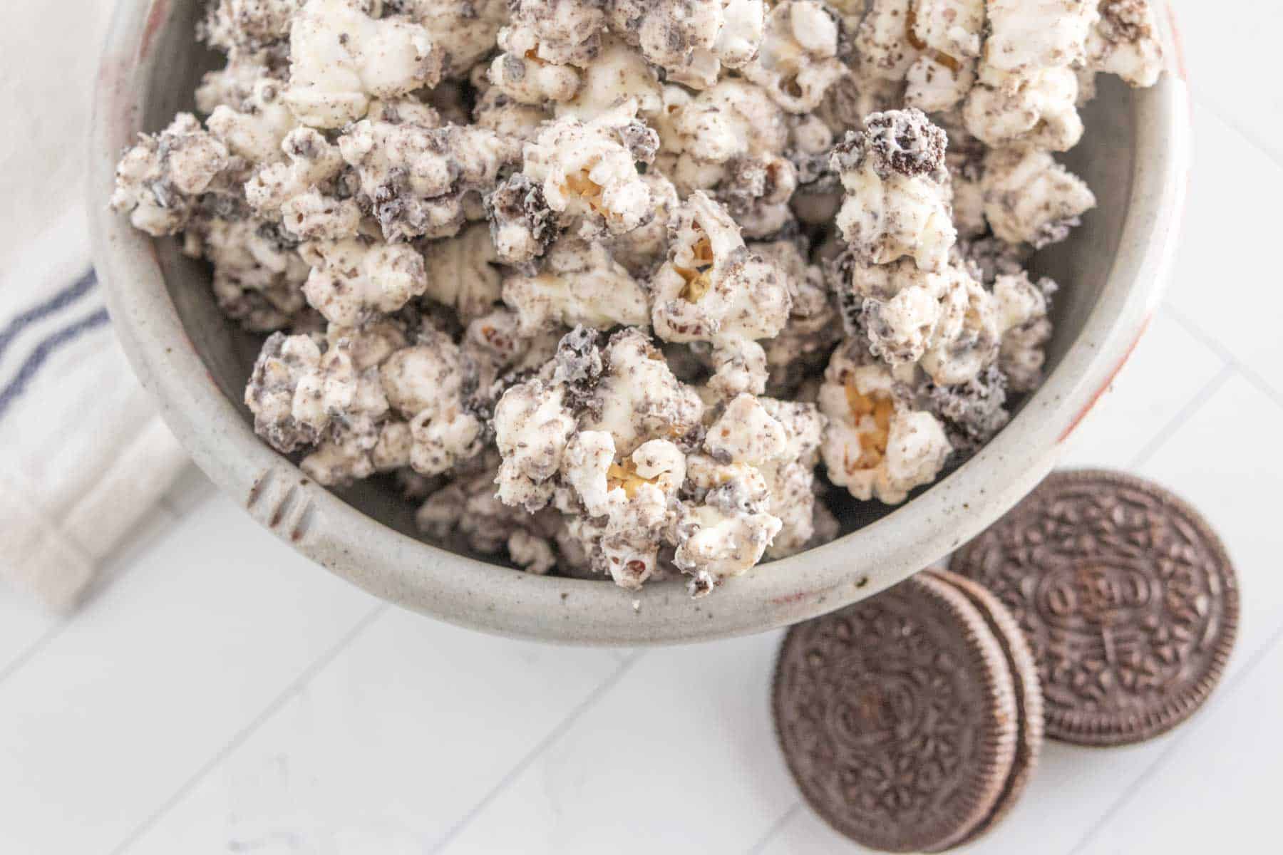 A bowl of popcorn coated in crushed cookies and cream with two chocolate sandwich cookies next to the bowl on a white surface.