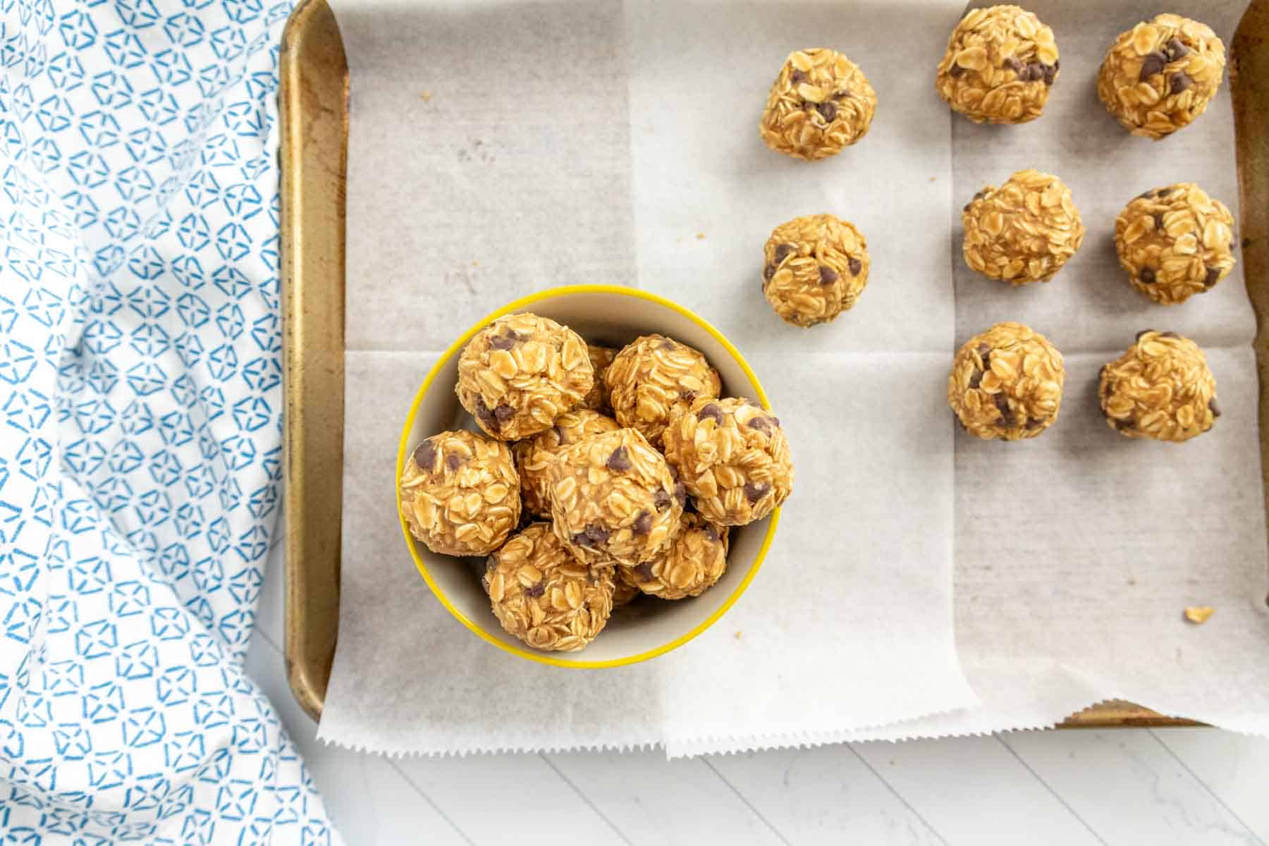 A bowl of oatmeal energy bites with chocolate chips sits on a tray lined with parchment paper. More energy bites are arranged on the tray next to the bowl.