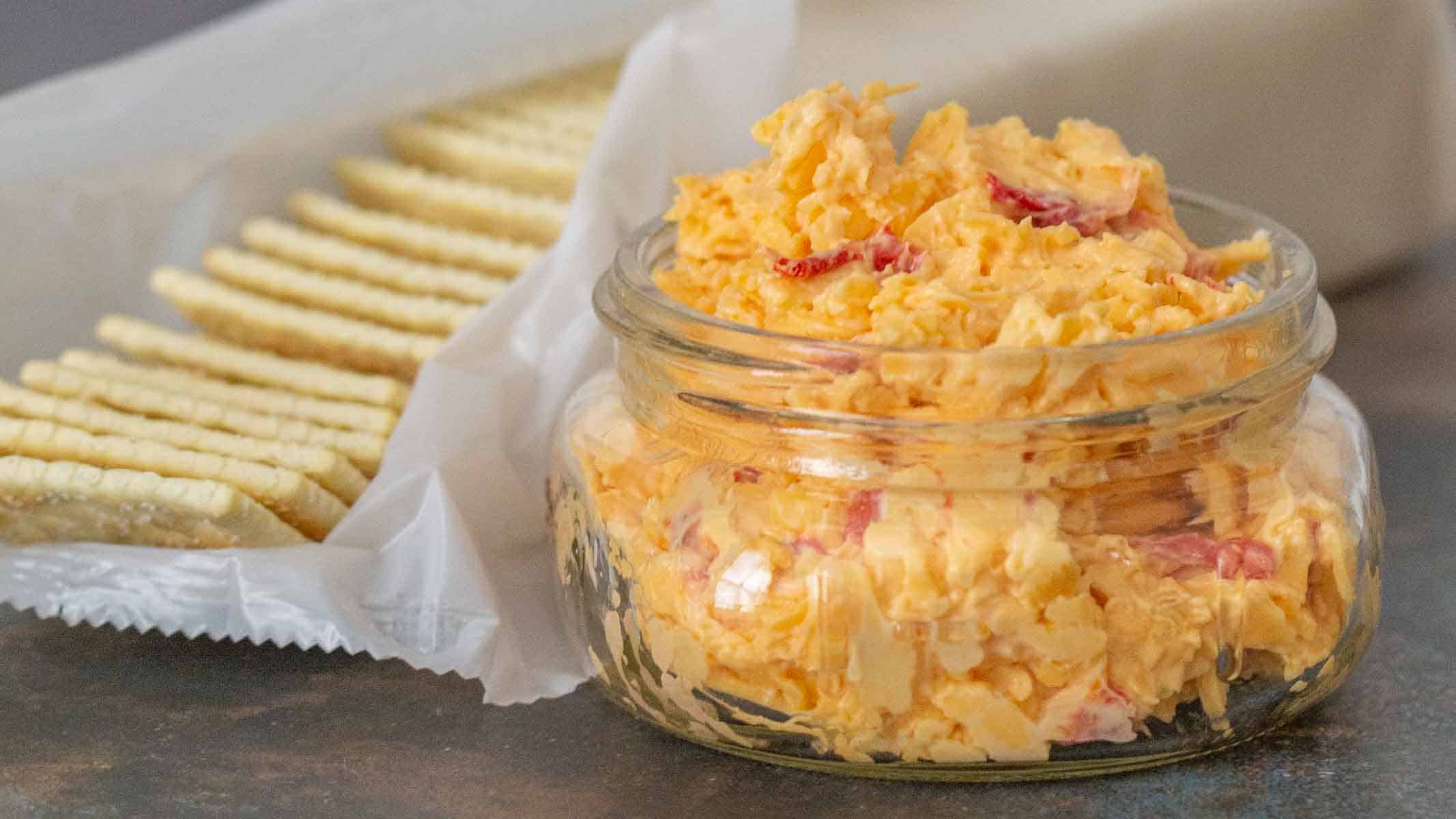A small glass jar filled with pimento cheese spread sits next to a stack of rectangular crackers on wax paper.
