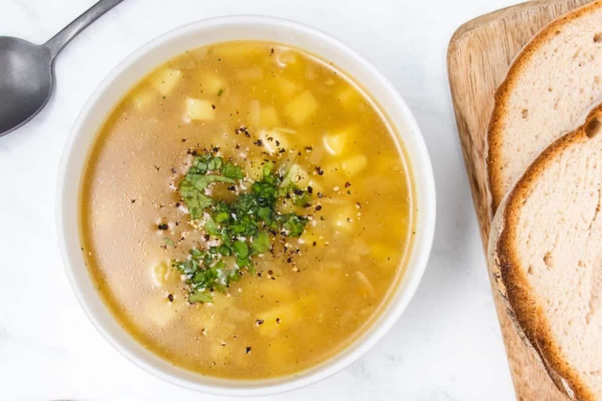 A bowl of clear soup with diced potatoes and herbs, next to two slices of bread on a wooden board and a silver spoon.