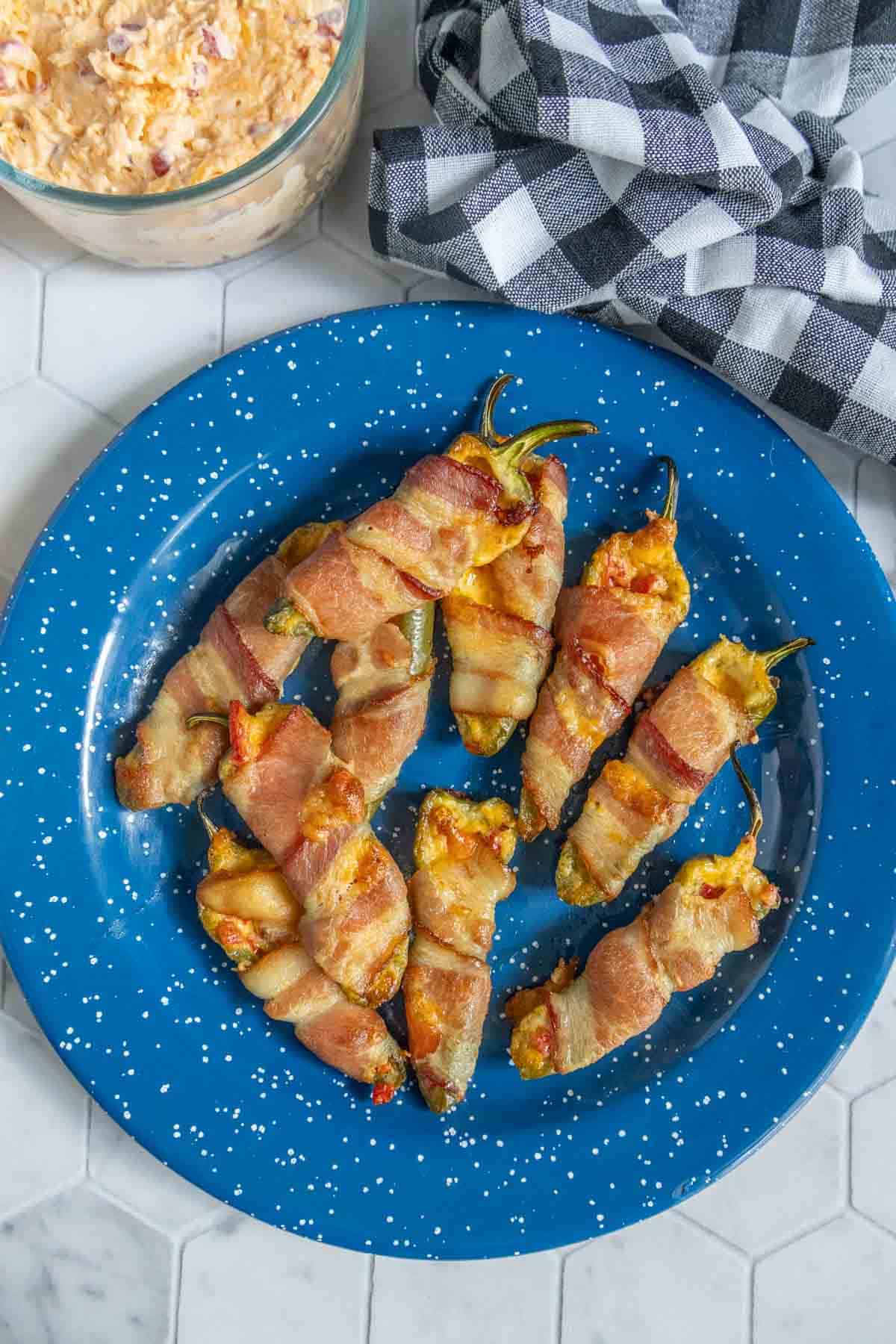 A blue plate with seven bacon-wrapped stuffed jalapeños, with a checkered cloth and a bowl of filling nearby on a white tiled surface.