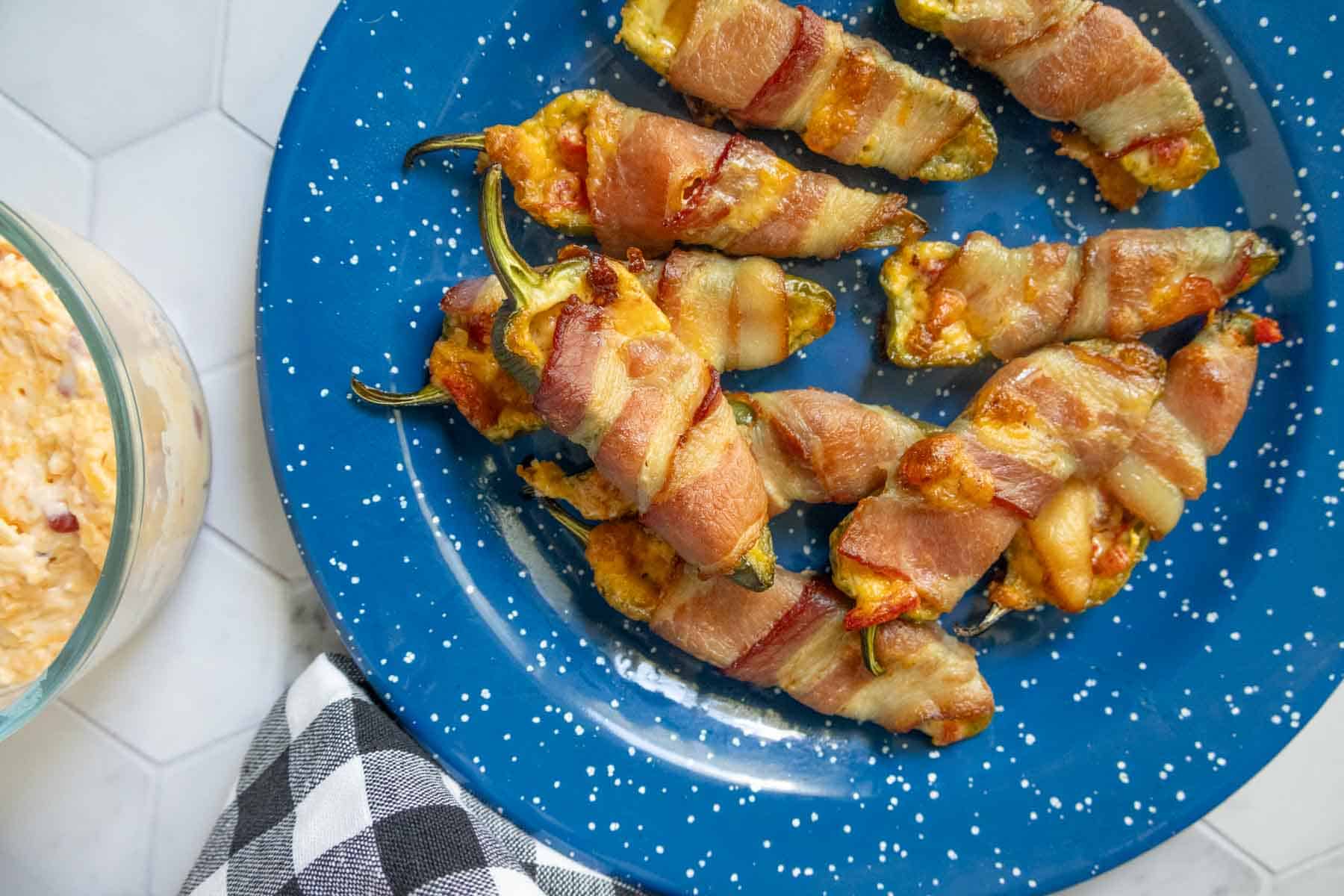 A blue plate with several bacon-wrapped jalapeño poppers, some with visible stems, next to a glass bowl and a black and white checkered napkin.