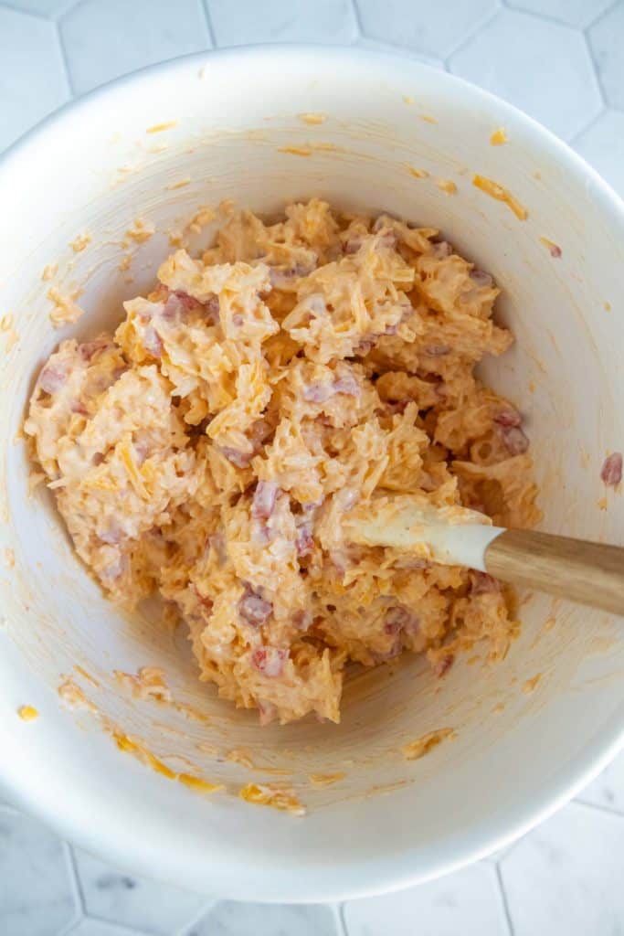 A white mixing bowl containing a mixture of shredded cheese, mayonnaise, and diced ham or bacon, with a spatula resting inside.
